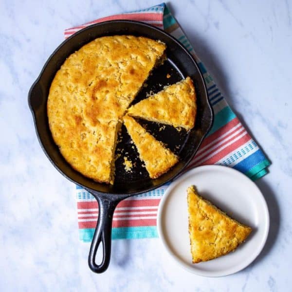 Cast iron skillet with cornbread sliced up. One slice is on a white plate.